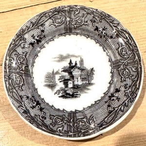 May include: A round ceramic saucer featuring a black and white transferware pattern. The center displays a detailed landscape with a small building, while the wide rim is decorated with intricate scrolling floral motifs and linear textures.