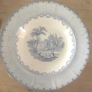 May include: A round ceramic plate featuring a cream-colored base with a blue transferware pattern. The center depicts a landscape with trees, a gazebo, and figures. The wide rim has a textured lattice design with a decorative floral border.
