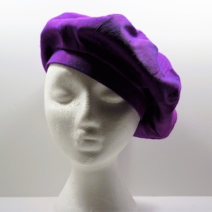 Peut inclure: Un béret en soie violet avec une texture douce et plissée. Le béret est porté sur une tête de mannequin blanche.