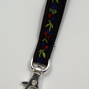 May include: Black lanyard with a floral pattern in red, blue, and green. The lanyard has a silver-coloured metal swivel clasp. The floral design is woven into the black fabric.