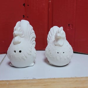 May include: A pair of white ceramic chicken-shaped salt and pepper shakers. Each shaker features a textured plumage design and two small holes on the front. A ruler visible in the background indicates a height of approximately 3 to 4 inches.