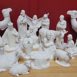 May include: A collection of unpainted white ceramic nativity figurines including two camels, three wise figures, an angel, a central holy family group, a donkey, a sheep, and a lamb, all featuring detailed molded textures and smooth matte surfaces.