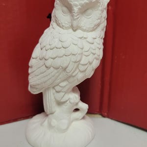 May include: A white ceramic owl figurine perched on a textured branch base. The owl features detailed feather carvings and closed eyes. A red measuring tool is visible in the background, indicating the figure stands approximately 6 inches tall.