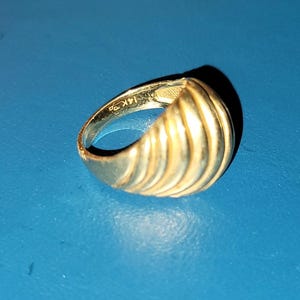 Puede incluir: Un anillo dorado con un dise&ntilde;o acanalado sobre una superficie azul. El anillo est&aacute; marcado "14K" en el interior. El dise&ntilde;o del anillo presenta crestas verticales, creando una apariencia texturizada. El anillo es una joya.