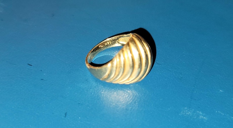 Puede incluir: Un anillo dorado con un dise&ntilde;o acanalado sobre una superficie azul. El anillo est&aacute; marcado "14K" en el interior. El dise&ntilde;o del anillo presenta crestas verticales, creando una apariencia texturizada. El anillo es una joya.