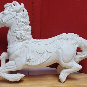 May include: A white ceramic carousel horse figurine featuring intricate floral carvings along the mane and saddle area. The horse is depicted in a galloping pose with a detailed bridle and a scalloped saddle blanket design.