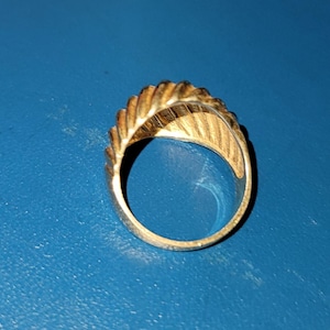 Puede incluir: Un anillo dorado con un dise&ntilde;o texturizado y acanalado en la parte superior y los lados. El anillo est&aacute; sobre un fondo azul, mostrando sus intrincados detalles y brillo met&aacute;lico. El dise&ntilde;o del anillo recuerda a una hoja o pluma.