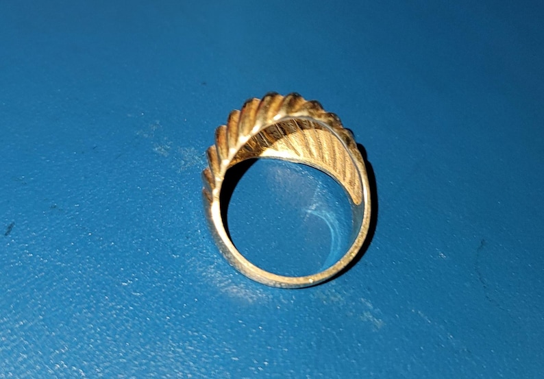 Puede incluir: Un anillo dorado con un dise&ntilde;o texturizado y acanalado en la parte superior y los lados. El anillo est&aacute; sobre un fondo azul, mostrando sus intrincados detalles y brillo met&aacute;lico. El dise&ntilde;o del anillo recuerda a una hoja o pluma.