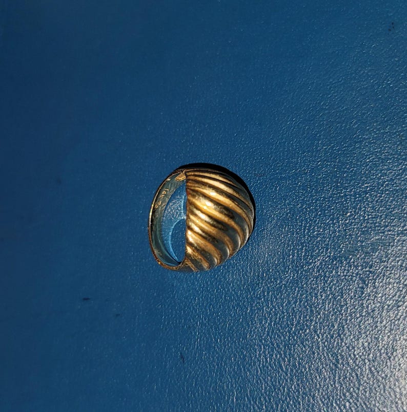 Puede incluir: Un anillo dorado con un dise&ntilde;o acanalado en forma de media esfera. El anillo est&aacute; sobre un fondo azul. La superficie del anillo refleja la luz, resaltando las crestas y curvas del dise&ntilde;o. El anillo parece ser de metal.