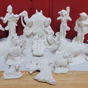 May include: A collection of unpainted white ceramic nativity figurines including figures of people, a camel, sheep, and a small angel. The set features various characters standing and kneeling, with a central baby in a manger, all in matte white.