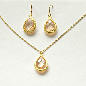 Puede incluir: Un conjunto de joyas en tono dorado que incluye un collar y pendientes a juego. El conjunto incluye un collar con colgante y dos pendientes colgantes, cada uno con una piedra preciosa en forma de lágrima de color melocotón engastada en un marco dorado texturizado.