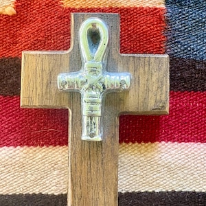 May include: A wooden cross with a silver ankh symbol attached. The cross is made of light brown wood with visible grain. The ankh is a silver color and is attached to the center of the cross. The background is a colorful woven textile.