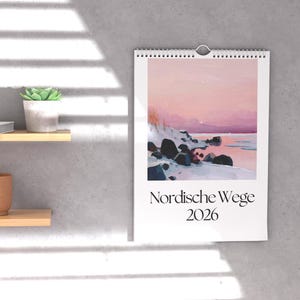 May include: A wall calendar with a winter landscape painting featuring a pink and purple sky, snow-covered rocks, and a body of water. The text "Nordische Wege 2026" is printed in black at the bottom.