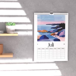 May include: A wall calendar with a landscape painting of a coastal scene, featuring blue water, colorful rocks, and a pink and blue sky. The calendar is labeled "Juli" and includes a monthly grid.