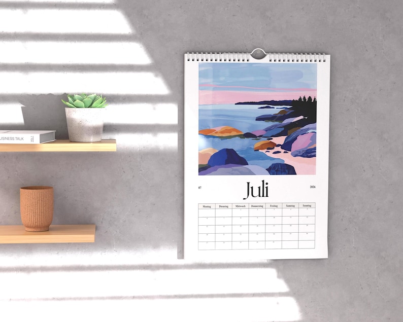 May include: A wall calendar with a landscape painting of a coastal scene, featuring blue water, colorful rocks, and a pink and blue sky. The calendar is labeled "Juli" and includes a monthly grid.