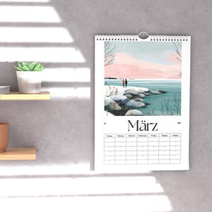 May include: A wall calendar with a watercolor landscape scene of two figures on a rocky shore, with a pink and blue sky. The word "M&auml;rz" is printed at the bottom. The calendar is hanging on a gray wall.