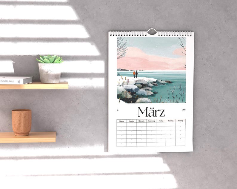 May include: A wall calendar with a watercolor landscape scene of two figures on a rocky shore, with a pink and blue sky. The word "M&auml;rz" is printed at the bottom. The calendar is hanging on a gray wall.
