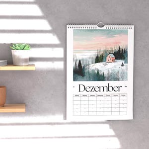 May include: A wall calendar with a winter landscape scene featuring a pink house, snow-covered hills, and evergreen trees. The word "Dezember" is printed at the bottom of the image. The calendar is hanging on a gray wall.