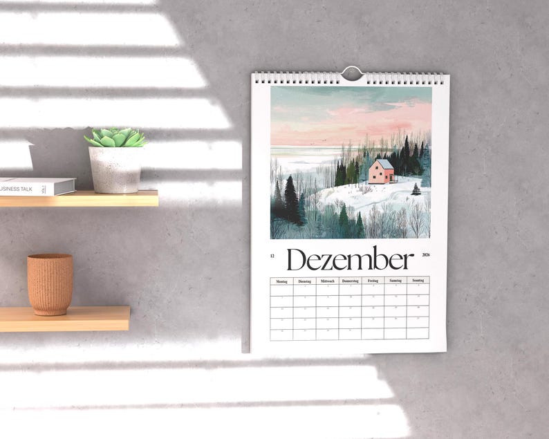 May include: A wall calendar with a winter landscape scene featuring a pink house, snow-covered hills, and evergreen trees. The word "Dezember" is printed at the bottom of the image. The calendar is hanging on a gray wall.