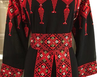 May include: A black dress with red and green embroidery. The dress has long sleeves and a round neckline. The embroidery is in a geometric pattern and includes small beads.