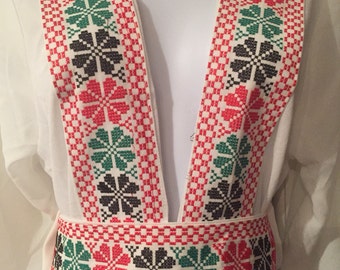 May include: White linen tunic with a deep V-neckline and a red, green, and black embroidered trim. The trim features a repeating pattern of flowers and geometric shapes. The tunic has a matching embroidered belt.