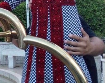 May include: A young child wearing a traditional embroidered dress with a red and white checkered pattern. The dress features intricate embroidery in red thread.