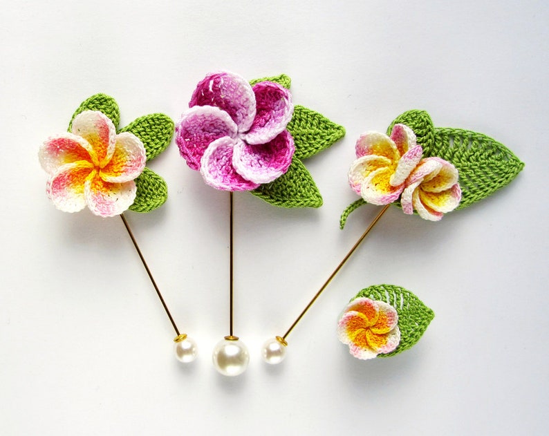 May include: Three crocheted flower pins with pearl heads. The flowers are pink, yellow, and white with green leaves.