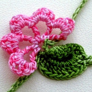 Puede incluir: Primer plano de una flor de ganchillo con p&eacute;talos rosas y una hoja verde. La flor est&aacute; unida a un tallo verde. El trabajo de ganchillo es detallado, mostrando la textura del hilo. Un art&iacute;culo de artesan&iacute;a floral.