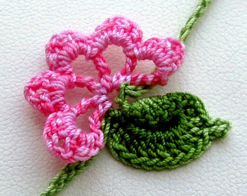 Puede incluir: Primer plano de una flor de ganchillo con p&eacute;talos rosas y una hoja verde. La flor est&aacute; unida a un tallo verde. El trabajo de ganchillo es detallado, mostrando la textura del hilo. Un art&iacute;culo de artesan&iacute;a floral.