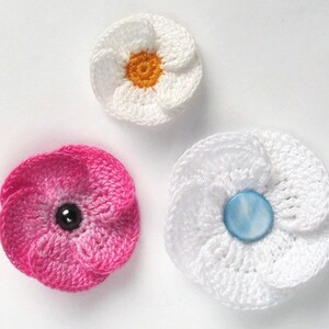 May include: Three crocheted flowers with different colored centers. The first flower is white with a yellow center. The second flower is pink with a black center. The third flower is white with a blue center.