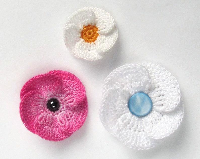 May include: Three crocheted flowers with different colored centers. The first flower is white with a yellow center. The second flower is pink with a black center. The third flower is white with a blue center.