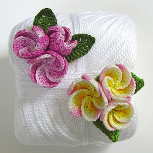 May include: Two crocheted flower embellishments on a white ball of yarn. One flower is pink and white, the other is yellow and white.
