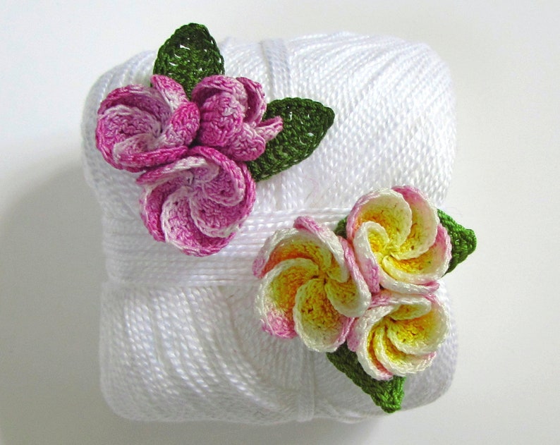 May include: Two crocheted flower embellishments on a white ball of yarn. One flower is pink and white, the other is yellow and white.