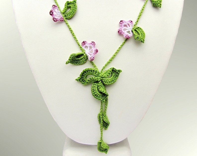Puede incluir: Un collar de ganchillo hecho a mano con un dise&ntilde;o floral. El collar tiene vides verdes con hojas verdes y flores rosas y blancas. El collar se exhibe en un maniqu&iacute; blanco.