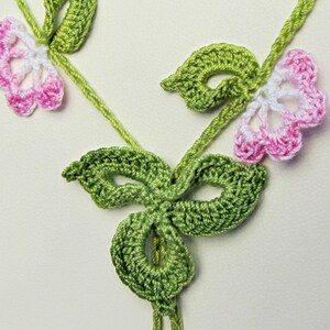 Puede incluir: Primer plano de un dise&ntilde;o floral de ganchillo hecho a mano. La pieza presenta hojas y tallos verdes, con delicadas flores en blanco y rosa. El trabajo de ganchillo es detallado e intrincado, mostrando una est&eacute;tica artesanal.