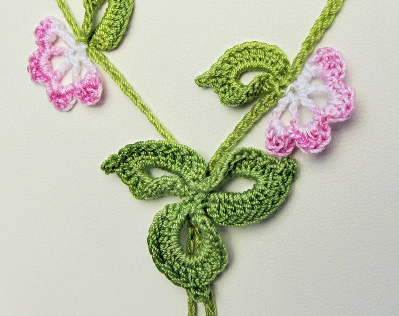 Puede incluir: Primer plano de un dise&ntilde;o floral de ganchillo hecho a mano. La pieza presenta hojas y tallos verdes, con delicadas flores en blanco y rosa. El trabajo de ganchillo es detallado e intrincado, mostrando una est&eacute;tica artesanal.