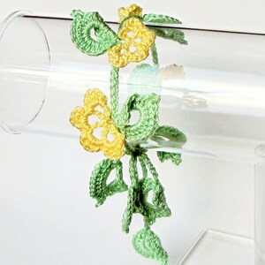 Puede incluir: Un brazalete de ganchillo hecho a mano con dise&ntilde;os florales amarillos y verdes. El brazalete est&aacute; hecho de hilo delicado, con flores amarillas y hojas y tallos verdes. El brazalete se exhibe en un soporte cil&iacute;ndrico transparente.