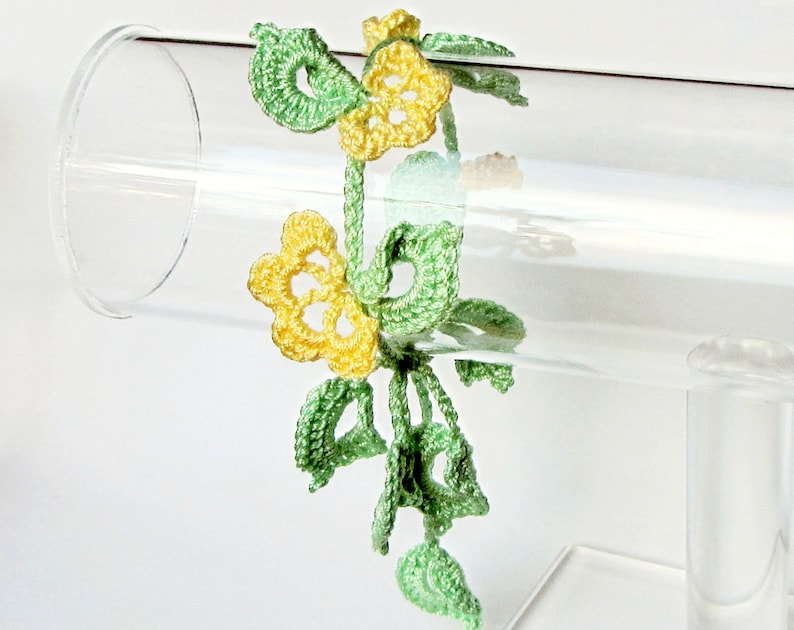 Puede incluir: Un brazalete de ganchillo hecho a mano con dise&ntilde;os florales amarillos y verdes. El brazalete est&aacute; hecho de hilo delicado, con flores amarillas y hojas y tallos verdes. El brazalete se exhibe en un soporte cil&iacute;ndrico transparente.