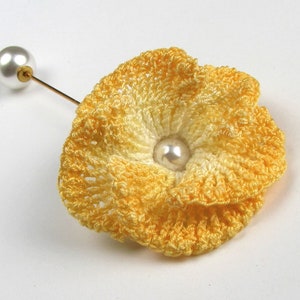 May include: A yellow crocheted flower brooch with a white pearl in the center. The brooch is attached to a gold pin.