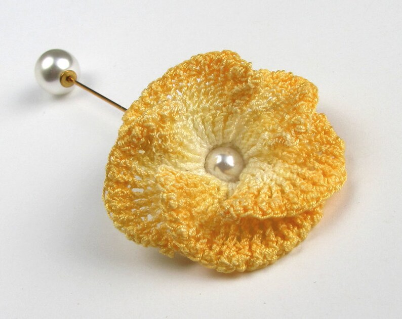 May include: A yellow crocheted flower brooch with a white pearl in the center. The brooch is attached to a gold pin.