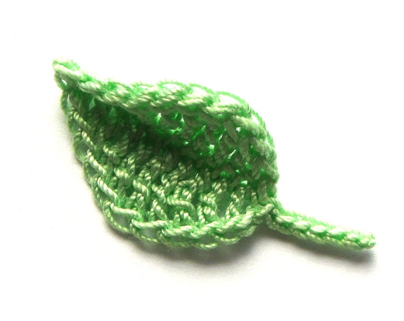 May include: A green crocheted leaf with a pointed tip and a stem. The leaf is made with a delicate stitch pattern.