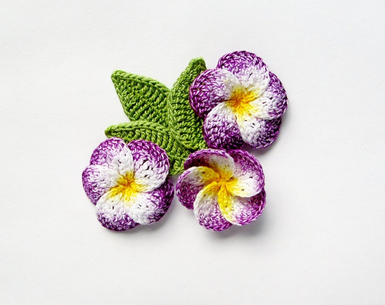 May include: Three crocheted purple and white flowers with yellow centers and a green crocheted leaf.