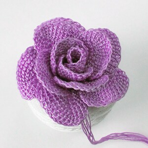 May include: A crocheted purple rose with a layered design. The rose is sitting on a white surface.