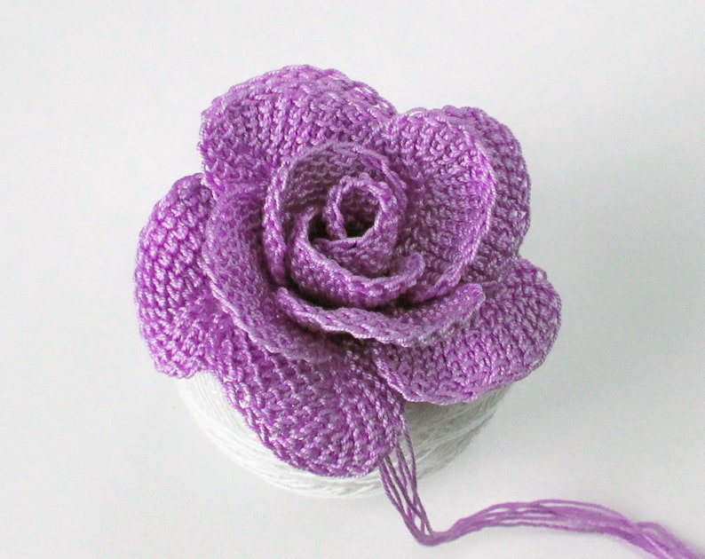 May include: A crocheted purple rose with a layered design. The rose is sitting on a white surface.