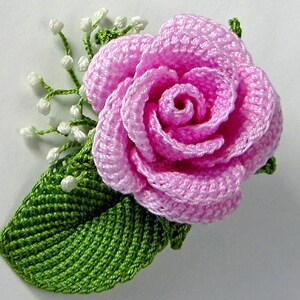 May include: A pink crocheted rose with green crocheted leaves and white crocheted flower buds.