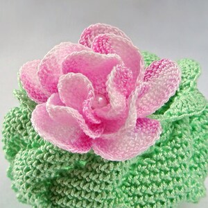 May include: A crocheted flower with pink petals and a green crocheted base. The flower has a pearl in the center.