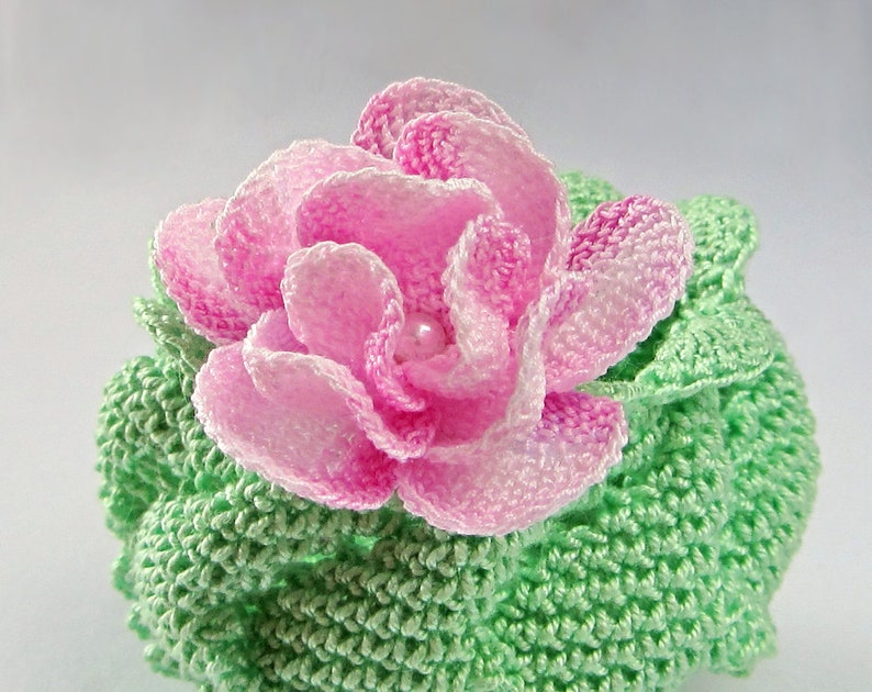 May include: A crocheted flower with pink petals and a green crocheted base. The flower has a pearl in the center.
