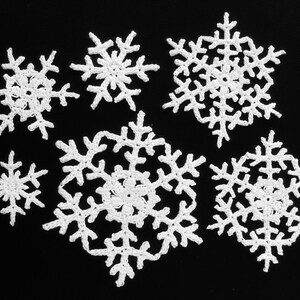 以下が含まれることがあります： 黒い背景に、さまざまな大きさの白いクロシェの雪の結晶が7つ並んでいます。