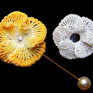 May include: Two crocheted flower brooches, one yellow and white, the other white. The yellow and white brooch has a pearl in the center and is attached to a gold pin with a pearl on the end.