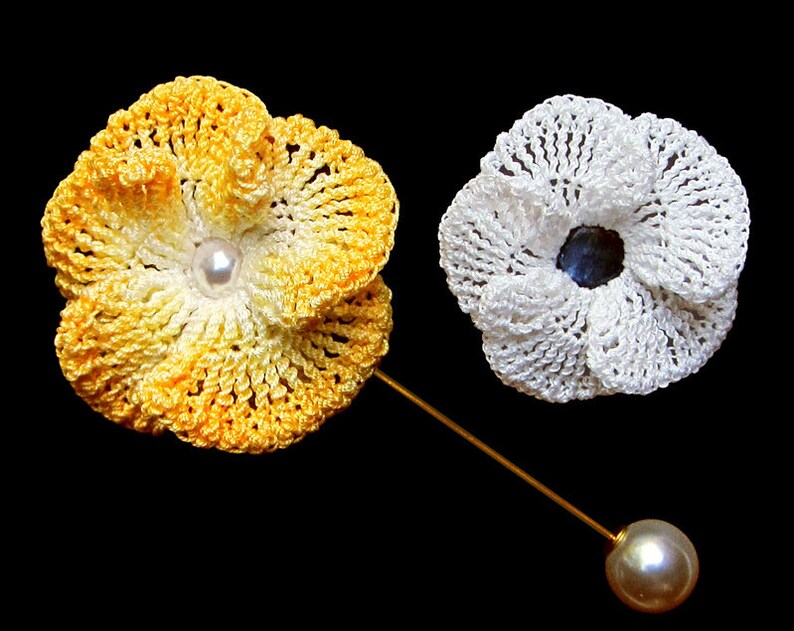 May include: Two crocheted flower brooches, one yellow and white, the other white. The yellow and white brooch has a pearl in the center and is attached to a gold pin with a pearl on the end.